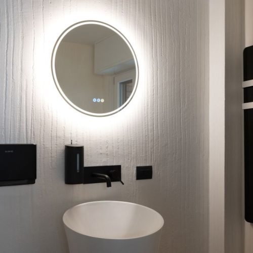 Alt text: bagno moderno con lavabo scultoreo, specchio rotondo retroilluminato e accessori neri opachi